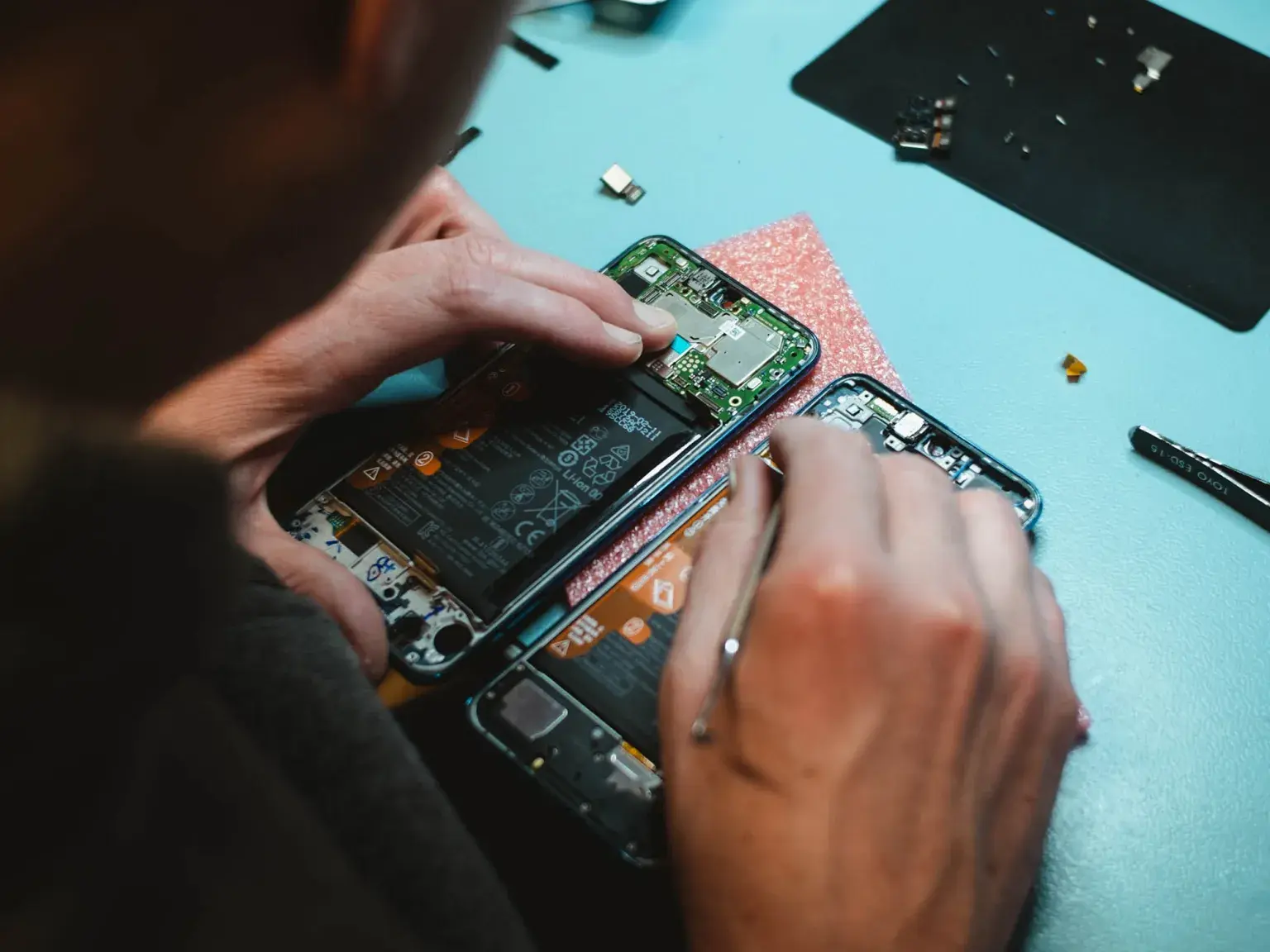 Man working on repair of a cell phone with its screen dissasembled at a blue table; using secondary but identical phone on left handside as guide for the repair.
